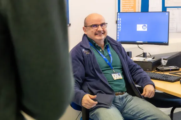 A male adviser sitting at his desktop engaging in conversation with a colleague and smiling
