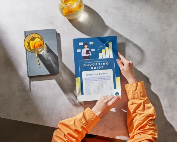 An aerial shot of a person holding a budgeting booklet guide with a diary and drink on the table to the left of the person. 