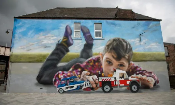 A mural on the side of a house of a young boy playing with toy cars