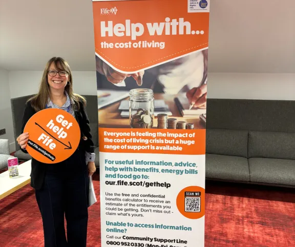 A female standing in front of a help with the cost of living pull up orange banner, holding a cardboard cutout of the get help fife logo.